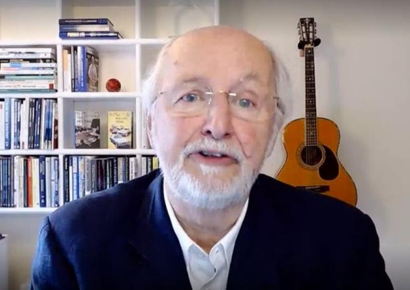 Image of Charles Jennings in his home office with bookshelf behind him and a guitar propped against wall. 