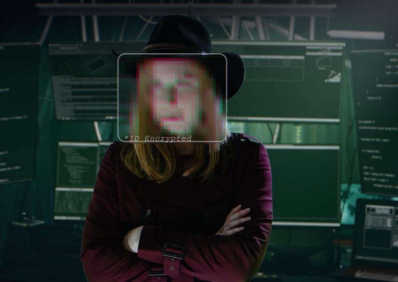 Photo of a person with face pixelated standing in a darkened room in front of a bank of monitors and computer equipment.