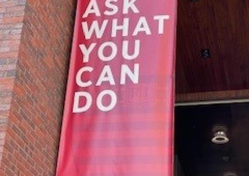 Photo of hanging wall sign affixed to a brick wall saying, ‘Ask what you can do’.