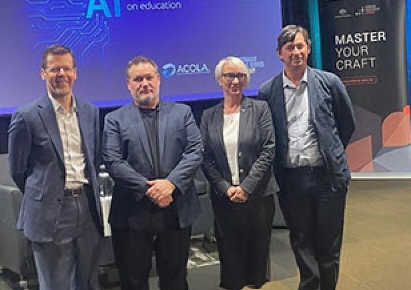 Photo taken of four speakers inside the Department of Education seminar room with stage and organisational banners behind them. 