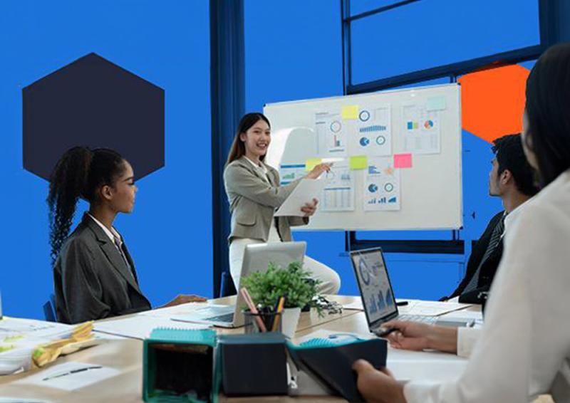 Three people sitting around a desk with a person pointing to graphs on a whiteboard.