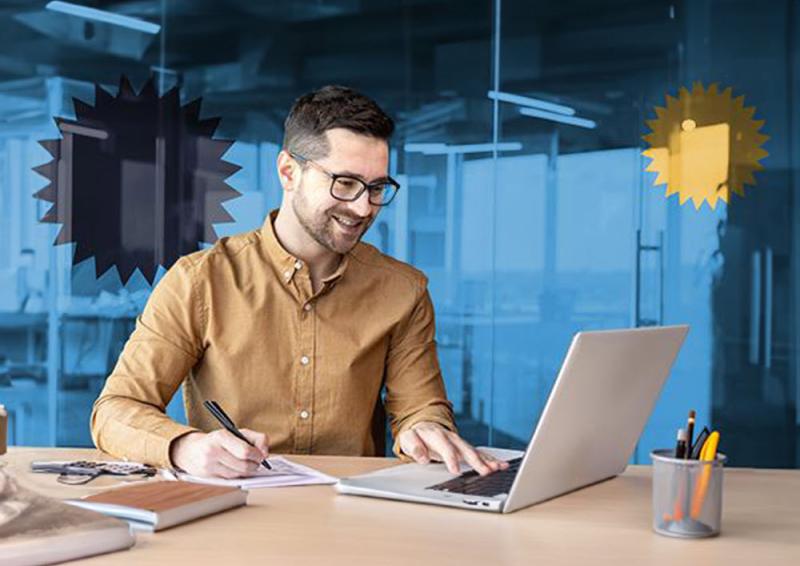 Person sitting at a desk working on a laptop and writing on a piece of paper