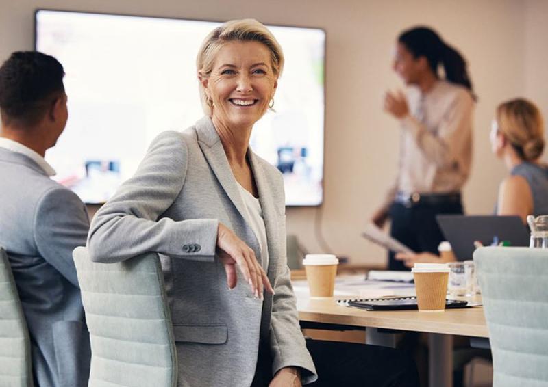 Photo of people sitting around a meeting desk look at a presentation, with a person smiling and looking directly in the camera