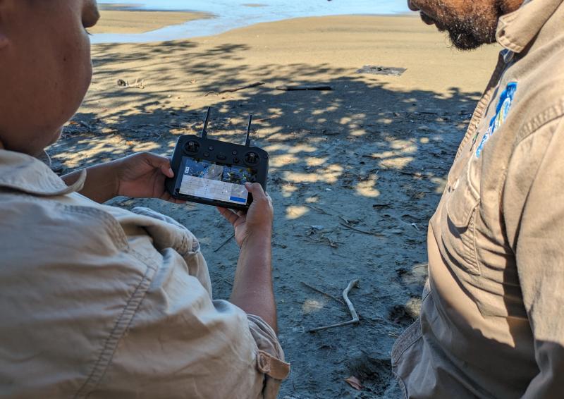 Image of two First Nations men standing on Larrakia land piloting a drone through a handheld device.