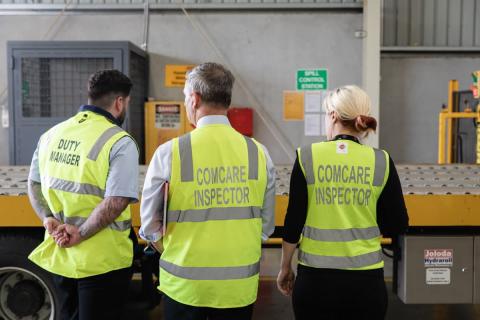 A photo featuring three people’s backs wearing hi visibility vests looking at a work area inside an industrial building.