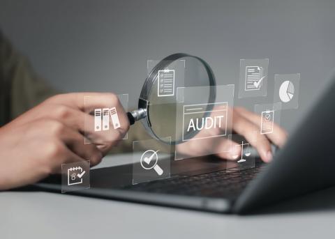 Illustrative image of person’s hand holding magnifying glass in front of a laptop with some floating graphics of checklist, word AUDIT, pie chart and folders