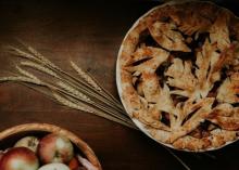 Apple pie in a dish atop a table also with a sheaf of wheat