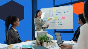 Three people sitting around a desk with a person pointing to graphs on a whiteboard.