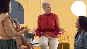 Person sitting on the edge of a desk, hands in an open palm gesture talking to three people.