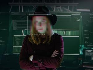 Photo of a person with face pixelated standing in a darkened room in front of a bank of monitors and computer equipment.