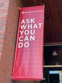 Photo of hanging wall sign affixed to a brick wall saying, ‘Ask what you can do’.