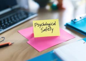 Photo of a desk with post it notes on top along with keyboard and folder. Wording is, “Psychological Safety’. 